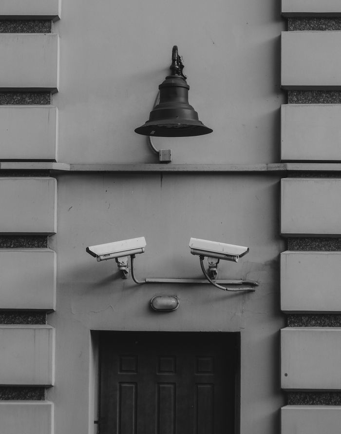 Black and white image of a building facade with two security cameras and a wall sconce.